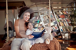 A young mother and her little daughter are posing while reading a book sitting on the swing at home together. Family, together,