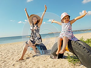 A young mother with her little daughter raises her hands joyfully up sitting near the sea on half-buried tires in the