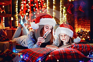 Young mother with her little daughter reading a book while sitting under decorated Christmas tree on the floor