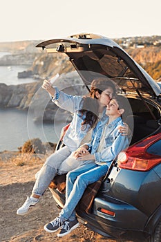 Young mother and her teenage boy in denim fashion clothes sitting in open car trunk, hugging and make selfie