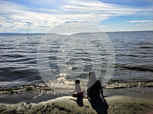 A young mother and her toddler daughter sitting together alone on a sandy beach watching the sparkling lake water