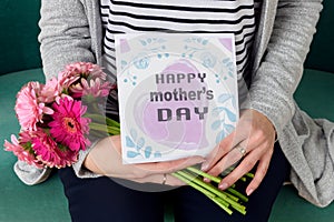 Young mother holding bouquet of gerbera daisies and mother`s day greeting card sitting on a couch.