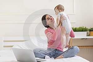 Young mother holding newborn baby while sitting on bed with laptop computer, spending time together at home