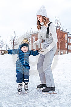 Young mother holds hand of son for boy for 2-3 years, ice skating rink winter in city park. Concept first lesson skating