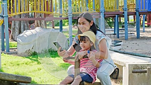 Young mother holds a smartphone to take pictures of her daughter playing seesaw in an outdoor playground. Happy child girls with m