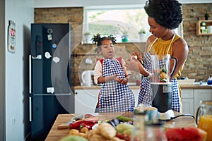 Young mother in the home kitchen with her little daughter, cutting up vegetables and tasting cucumber slices, girl acting silly