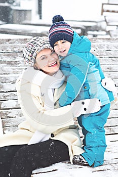 Young mother hugging with toddler boy sitting on bench in winter park. Woman playing and hugging with cute baby son