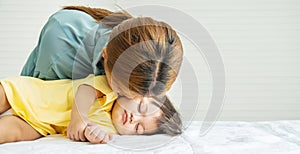 Young mother is kissing her cute little baby who is sleeping in bed at home, Mom and daughter relax in a white bedroom.