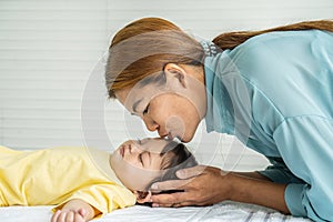 Young mother is kissing her cute little baby who is sleeping in bed at home, Mom and daughter relax in a white bedroom.