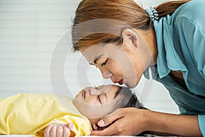 Young mother is kissing her cute little baby who is sleeping in bed at home, Mom and daughter relax in a white bedroom.