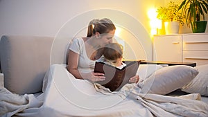 Young mother kissing her toddler son reading book in bed at night