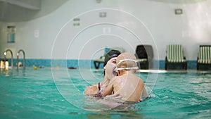 Young mother lifting her little boy in protective glasses from the water while teaching him how to swim in the swimming