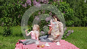 Young mother and litlle daughter on a spring picnic next to a blossoming lilac.