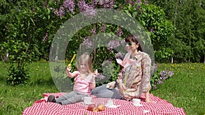 Young mother and litlle daughter on a spring picnic next to a blossoming lilac.