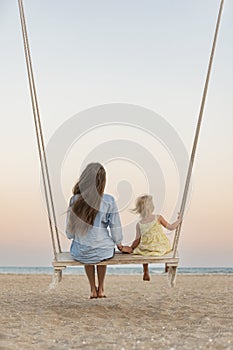 Young mother and little blond girl sit on swing on the beach during sunset. Mom and daughter on sea, magic sunset lights