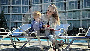 A young mother with a little blond son playing on the lawn and reading a book near the pool.