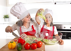 Young mother and little daughter at house kitchen preparing salad for lunch wearing apron and cook hat