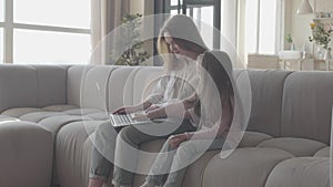 A young mother and little daughter with long hair sitting together on the sofa in the living room typing on a laptop