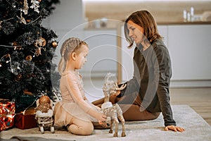 A young mother and little daughter play sitting near the Christmas tree.