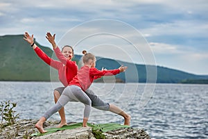 Young mother and little daughter practicing balancing yoga pose on the rock near river.