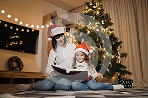Young mother and little daughter in santa hat reading fairy tale book girl sitting near tree