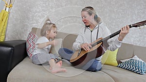 A young mother and a little girl learn to play acoustic guitar sitting at home on the couch.