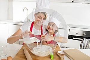 Young mother and little sweet daughter in cook hat and apron cooking together baking at home kitchen