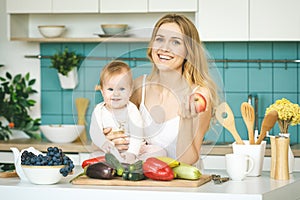 Young mother looking at camera and smiling, cooking and playing with her baby daughter in a kitchen. Healthy food concept