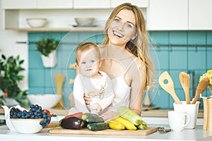 Young mother looking at camera and smiling, cooking and playing with her baby daughter in a kitchen setting. Healthy food