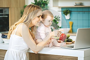 Young mother looking at camera and smiling, cooking and playing with her baby daughter in a modern kitchen. Using laptop. Healthy