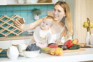 Young mother looking at camera and smiling, cooking and playing with her baby daughter in a modern kitchen. Using phone, making