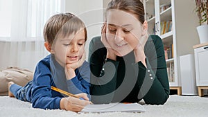 Young mother looking at her little son doing homework on carpet in living room. Concept of child education, development