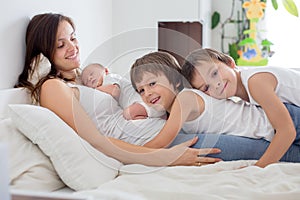 Young mother lying in bed with her newborn baby boy and his older brothers, holding them in her arms and smiling