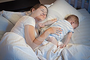 Young mother lying on bed at night next to her sleeping baby boy