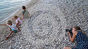 Young mother lying on pebble beach in the evening to take photos of her three children