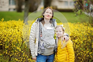 Young mother with a newborn in a baby carrier hugging her older daughter. Cute young child having fun with her mom and sibling