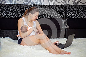 Young mother with a newborn in her arms works at home on a laptop, sitting on a sofa