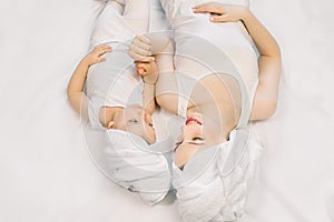 Young mother playing with her baby girl on bed at home. Mom and cute daughter in white towels on heads, lying together