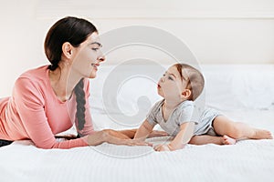 Young mother playing with her cute baby, talking and spending time together, lying on bed in bedroom at home