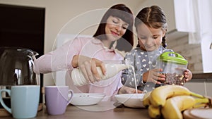Young mother pouring milk into daughters bows of cornflakes for breakfast in light modern kitchen.