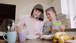 Young mother pouring milk into daughters bows of cornflakes for breakfast in light modern kitchen.