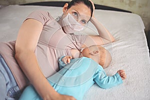 Young mother in protective face mask with newborn cute infant baby breastfeeds him with breast milk