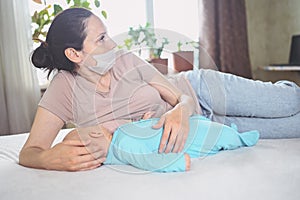 Young mother in protective face mask with newborn cute infant baby breastfeeds him with breast milk