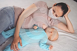 Young mother in protective face mask with newborn cute infant baby breastfeeds him with breast milk