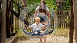 Young mother pushing her baby son swinging in rope nest swing in park. Kids playing outdoors, children having fun, summer vacation