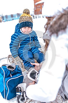 A young mother puts on skates, helps a little boy of 2-4 years old child. In winter, on the rink in city. Rest on the