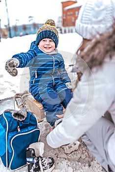 Young mother puts skates in winter public skating rink. The little boy happy smiling shows his hand to the side, in blue