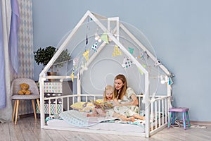 Young mother reading a book with her 3-year-old toddler girl while sitting in cute house bed