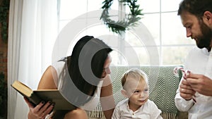 Young mother reading book for her son and husband. Family reading Christmas story while sitting by Christmas tree. Happy