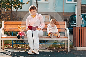 Young mother is reading a book sitting on a bench. A child is sitting nearby and watching cartoons on a smartphone